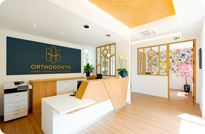 Accueil et salle d'attente du cabinet d'orthodontie des Docteurs Davy et Alam, avec le logo sur le mur, bureau en bois clair, décoration florale et grande baie vitrée
