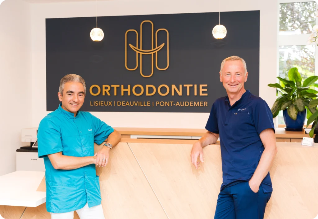 Les Drs Jean-Luc Davy et Rafeh Alam dans leur cabinet d'orthodontie à l'accueil, avec l'enseigne Orthodontie Lisieux Deauville Pont-Audemer en arrière-plan